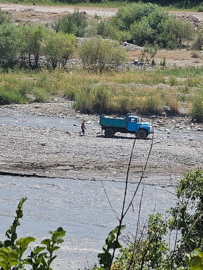 A blue truck is parked in a shallow, rocky riverbed. A single person, wearing dark shorts and no shirt, walks away from the truck towards the left of the frame. The riverbed is composed of small grey and brown stones, with some areas of darker, wet sand. The background shows a steep, overgrown bank with various types of green vegetation and trees. The sky is not visible. The scene appears to be a remote, natural setting.