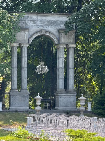 The image depicts an outdoor setting, likely a park or garden, featuring a classical-style stone archway with Corinthian columns. A crystal chandelier hangs from the arch, suspended by a chain. In the foreground, rows of clear acrylic chairs are arranged, suggesting an event or ceremony is about to take place. Two ornate stone urns flank the archway. In the background, lush green trees create a natural backdrop. The scene appears to be in Krubki-Górki, Poland, based on the provided location information. No people are visible in the image.