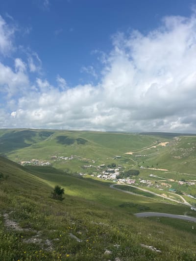 The image captures a vast, rolling green landscape under a bright blue sky with scattered white clouds. In the mid-ground, a small village is nestled in a valley, with buildings clustered together and a winding road passing through it. The road is paved and has white lines marking the lanes. Power lines and poles are visible along the road and in the distance. The foreground consists of a grassy slope dotted with small yellow wildflowers and some rocks. The overall scene depicts a rural, mountainous environment in the vicinity of Kichi-Balyk, Russia. There are no people visible in the image. The lighting suggests it is daytime, likely mid-morning or afternoon, with shadows indicating the sun is at an angle.