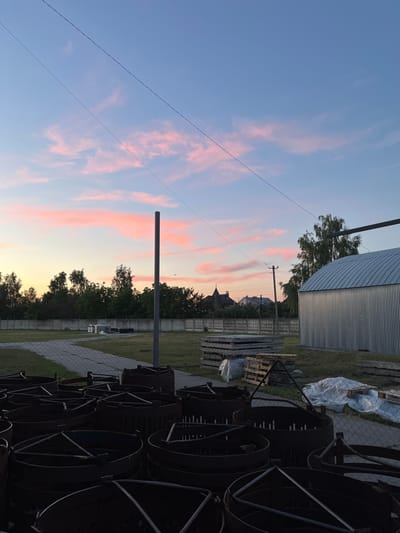 This is a vertically oriented, outdoor, landscape photograph taken at dusk in Zmiinets, Ukraine. The scene depicts a sky with wispy pink and blue clouds illuminated by the setting sun. In the foreground, to the right, is a stack of dark, cylindrical metal objects, possibly large spools or industrial components, casting long shadows. A chain-link fence runs along the edge of this area. Behind the fence, to the left, is a paved walkway and a patch of green grass. Further back, a line of trees forms a natural barrier, behind which are the rooftops of houses and a church steeple. To the far left of the image, a tall, thin metal pole stands vertically, and a power line stretches across the sky. To the far right, a corrugated metal building with a curved roof is visible, partially obscured by a tree. The overall atmosphere is calm and serene, characteristic of twilight. There is no visible text in the image.