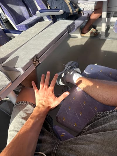 The image shows a close-up view of a person's legs and hand inside what appears to be a train carriage. The person is wearing grey sneakers with white socks and denim shorts. A metal tray table is extended in front of them. The seats in the train are upholstered in a purple fabric with a subtle yellow and white geometric pattern. Sunlight is streaming into the carriage, casting strong shadows on the seats and the person's legs, suggesting it is daytime. The angle of the light indicates the sun is relatively low in the sky, possibly morning or late afternoon. The person's hand is open, palm facing upwards, and positioned near the tray table, perhaps in a gesture of resting or preparing to use it. The overall impression is one of travel, with the sunlit interior of the train creating a warm and somewhat relaxed atmosphere. No text is visible in the image.
