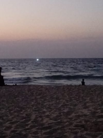The image depicts a twilight scene at a beach in Al-Mawasi, Palestinian Territories. The horizon line separates a graded sky, transitioning from light grey and pale lavender at the top to darker purplish-grey closer to the sea. The sea itself is dark, exhibiting visible waves breaking towards the shore. A distinct, bright white point of light is visible on the water's surface, approximately one-third from the left edge of the frame, near the horizon. The foreground consists of dark, textured sand. On the far left, a dark, indistinct silhouette, possibly a person or rock formation, is partially visible. To the right, near the water's edge, a small, dark silhouette of an individual, likely a child, sits facing the open sea. No specific actions or interactions beyond passive observation are discernable.