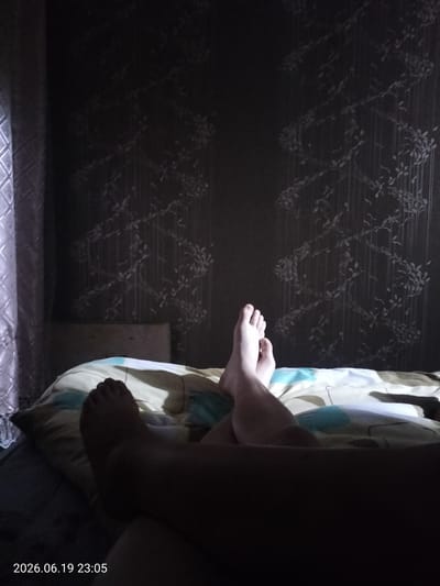 The image shows a person's legs and feet resting on a bed. The person appears to be lying down, possibly in a bedroom, as indicated by the presence of a pillow and bedding. The background features wallpaper with a subtle floral or vine pattern. A curtain or window covering is visible on the left side of the frame. The lighting in the room is dim, suggesting it is nighttime or the lights are turned down. The timestamp in the bottom left corner indicates the date and time as 2026.06.19 23:05. The overall mood is relaxed and private. No specific weather conditions are discernible. The location context provided is Ridder, Kazakhstan.