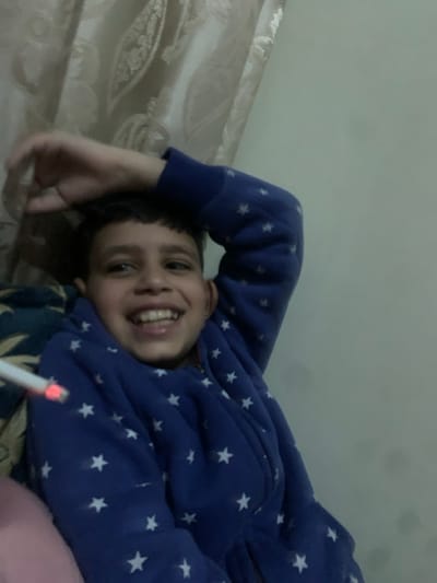 A child in a blue, star-patterned hoodie lies sideways on a bed with their head propped up by one hand. The child is smiling, facing the camera. Next to the child, at the bottom of the frame, is an object that appears to be a lit cigarette. The background includes a wall and patterned bedding.