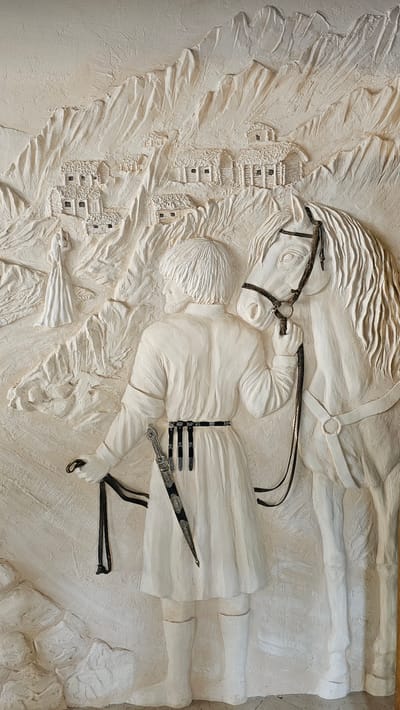 This is a relief sculpture made of light-colored material, depicting a scene set in a mountainous, village-like environment. In the foreground, a person stands facing away from the viewer, holding the reins of a white horse. The person is dressed in traditional attire, including a long robe, a patterned belt with what appears to be a dagger tucked into it, and high boots. The horse is saddled and bridled, its head turned slightly towards the person.

In the background, the mountainous terrain is textured to resemble rough rock faces and steep slopes. Clustered on these slopes are small, rustic buildings with thatched roofs, suggesting a remote village or settlement. To the far left of the frame, a solitary figure in a long, flowing dress stands on a path amidst the rocky landscape. The overall impression is a historical or folkloric scene, evoking a sense of tradition and a connection to nature. The location in the city of Красный Курган, Russia, is not depicted in the artwork itself, which presents a generic mountainous village scene.