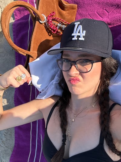 An overhead, high-angle image depicts a single individual lying on a beach in Manhattan Beach, United States, likely on a purple striped beach towel. The person, with long dark hair styled in two braids, wears a dark-colored baseball cap featuring a white "LA" logo, dark-framed eyeglasses, and a dark-colored bikini top. Their left arm is bent, with the hand partially clenched, displaying a gold-toned coin-like ring and a silver-toned ring with a cross motif, along with gold-toned bangles on the wrist. A layered necklace setup is visible, including a rosary-style necklace with a crucifix pendant and another chain with small charms. The individual is looking towards the camera with pursed lips. Behind their head, a white garment serves as a pillow. Adjacent to the individual, a brown suede-like tote bag rests on the towel, adorned with a red and white striped tassel keyring featuring multiple silver-colored charms, including a cross. Sand is visible in the foreground and around the towel, indicating a beach environment under direct sunlight.