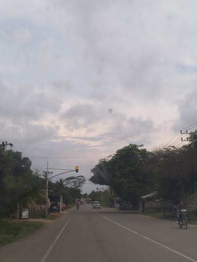 The image depicts a two-lane road in Cotentung, Indonesia, with a cloudy sky overhead. The road is bordered by trees and small buildings on both sides. Several vehicles are present, including a motorcycle with a rider in the foreground, a car and a truck in the distance, and a van on the right side of the road. A traffic light displaying yellow is visible on the left side of the road. The environment appears to be rural or semi-urban, with a focus on transportation infrastructure and natural elements.