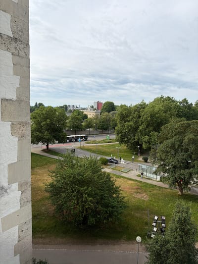 The image presents an elevated view of a city street in Ingolstadt, Germany, under an overcast sky. The foreground is dominated by a rugged, light-colored stone wall, possibly a parapet or balcony edge, featuring visible mortar lines and patches of white paint or plaster.
Looking down, a wide road with multiple lanes traverses the scene. On the right side of the road, a long, white and blue city bus is visible, possibly stopped at a bus stop. A paved sidewalk runs alongside the road, leading to a modern bus stop shelter with glass panels. Further down, another vehicle, possibly a white car, is parked. A person is standing near the bus stop shelter, and another individual on a bicycle is next to the bus. A distinct red bicycle lane is marked on the road.
Adjacent to the sidewalk and bus stop is a wide grassy area dotted with numerous large, lush green trees. Several streetlights are spaced along the pathway. Notably, a cluster of eight large spotlights mounted on a pole is positioned in the grassy area, suggesting an area used for events or sports.
On the left side of the road, there's another strip of grass and more trees, with residential or commercial buildings visible in the distance, forming a typical urban skyline. The sky above is bright but covered by a layer of streaky white clouds, indicating an overcast day. A green street sign and a blue circular sign are visible on the left side of the road, but any text on them is not legible from this distance. The overall impression is a bustling urban environment with a balance of infrastructure and green spaces.