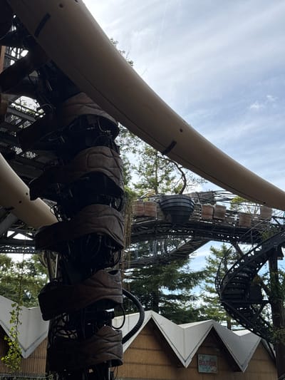 The image depicts a detailed view of the Chroniques de la Loire, a mechanical installation located in Nantes, France. The structure, designed to resemble a large, segmented creature or mechanical arm, is prominently featured. It is made of dark, textured material with visible pipes and wiring, giving it a complex, industrial aesthetic. The installation is elevated, with metal walkways and spiral staircases visible in the background, interwoven with lush green trees. The sky above is partially cloudy. In the lower part of the image, the peaked roofs of wooden structures are visible, with a sign for "BOUTIQUE" indicating commercial activity. No people are visible in the frame. The overall impression is one of intricate engineering integrated with a natural, tree-filled environment.
