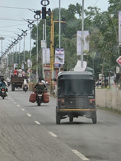 This is a street-level, eye-level shot of a road in Datala, India, with traffic moving. The scene is captured on an overcast day. In the foreground, a black auto-rickshaw is traveling away from the camera. The auto-rickshaw has an exhaust pipe that is emitting smoke. Behind the auto-rickshaw, a motorcycle is also traveling away from the camera. This motorcycle has two large, red, cylindrical objects attached to its rear. The road is paved with asphalt and marked with white dashed lines. On the left side of the road, there is a row of lamp posts and traffic lights. On the right side of the road, there is a concrete barrier with some vegetation growing behind it, and a sidewalk. There are several advertisements on poles along the roadside. Further in the background, a flatbed truck is visible, carrying several passengers. Other vehicles and people are also present on the road. The overall environment suggests a busy urban or semi-urban setting.