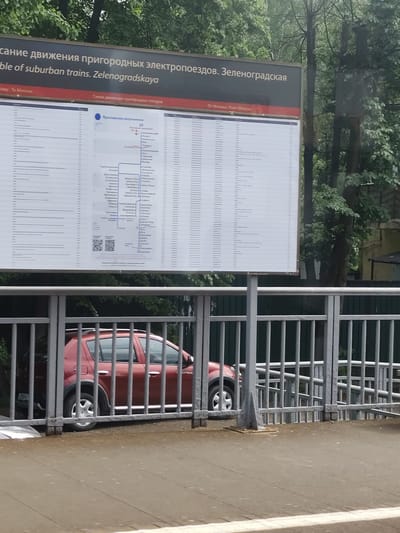 The image shows a timetable for suburban trains from Zelenogradskaya in Zelenogradskiy, Russia. The timetable is displayed on a large sign next to a metal fence. Behind the fence is a red car. The sign has text in Russian and English, indicating that it displays the schedule of suburban trains to and from Moscow. The timetable itself contains a diagram of the train routes and schedules. Trees and vegetation are visible in the background.