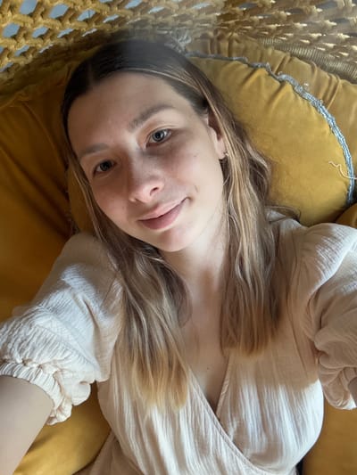 A woman with blonde hair and fair skin is lying down on a yellow cushion or pillow, wearing a white, long-sleeved, textured shirt. Part of a woven hammock or hanging chair is visible on the left side of the image. The background is blurred, but no elements indicate the location is Чёрное, Russia.