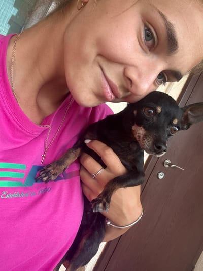 A young woman is holding a black and tan miniature pinscher dog. The woman has fair skin, light brown hair, and is wearing a pink t-shirt with a gold chain and a cross pendant. She has a silver ring on her finger and a silver bracelet on her wrist. The dog is looking up with brown eyes. The background is a dark brown door with silver hardware. It is not possible to determine from the image if the location is Kashin, Bulgaria.