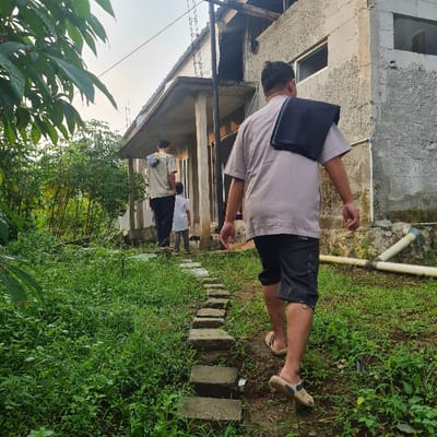 Three men are walking down a pathway of stone steps, covered in green foliage, in front of a residential building in Kecamatan Tamansari, Indonesia. The men are wearing casual attire, and one of the men is carrying a dark-colored cloth over his shoulder.  The environment includes a grey-colored building with visible piping, dense greenery, and green foliage. One man is wearing a white collared shirt and black pants. The man in the lead is wearing a grey short-sleeve shirt and black shorts. The building appears to be a single-story structure with windows and a door, and there is an open space behind the building. The men are walking towards the viewer, and the path leads away from the building. The scene is set against a backdrop of a clear sky.