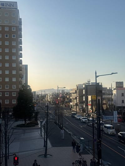 This image captures an urban street scene in Tokorozawa, Japan, under a clear, bright sky. The perspective is elevated, looking down a main thoroughfare that recedes into the distance towards distant, hazy mountains.

On the left, a tall, multi-story building, approximately 12 visible floors, dominates the foreground. It is constructed of brown/beige materials with numerous evenly spaced square windows. A white sign atop the building displays "横INN" (Yoko INN). Below this building, a paved pedestrian area features a circular planter with a small evergreen tree, surrounded by several bare deciduous trees. A pedestrian traffic light with a red "standing person" symbol is visible in the lower left, near two individuals in dark clothing. Further along the left sidewalk, a person in full white attire walks away from the viewer.

The central part of the image is occupied by a multi-lane asphalt road, extending into the background. The road appears to have at least two lanes of traffic moving away from the viewer on the right side, with a line of parked or slowly moving vehicles, predominantly white and silver sedans and SUVs. A white van is visible among them. The road is bordered by sidewalks on both sides, lined with numerous black utility poles and streetlights featuring decorative finials.

On the right side of the road, a dense collection of commercial buildings of varying heights and architectural styles line the street. Japanese signage is prevalent on these structures. A prominent white building further down the street displays "HOTEL LAIKA" and "Renewal." Closer to the foreground on the right, another building has signage, including "立呑みやきとり" (Tachinomi Yakitori). In the lower right foreground, several individuals are gathered on the sidewalk, some partially obscured by a streetlight pole, with at least one person accompanying a bicycle. A blue street sign with Japanese text is also visible in this area.

The overall lighting suggests either morning or late afternoon, with bright sunlight casting long shadows towards the viewer, particularly evident on the left sidewalk and from the vehicles. The environment appears cool, indicated by the bare branches of many trees.
