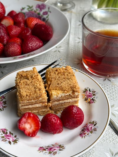The image shows a table setting in what appears to be a domestic environment in Ogresgals, Latvia. A plate decorated with floral patterns holds two pieces of cake with creamy layers, accompanied by three strawberries, one of which is cut in half. A fork rests on the plate beside the cake. To the left, a bowl overflows with fresh strawberries. Adjacent to the plate is a glass containing a dark liquid, possibly tea. The tablecloth has a subtle, patterned texture. The scene suggests a snack or dessert setting.
