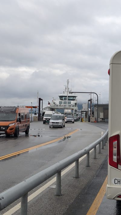 The image shows a ferry waiting to load vehicles at a dock in Halsa, Norway. The sky is overcast, and the road is wet, indicating recent rain. Several vehicles, including vans and cars, are lined up on the ferry ramp, waiting to board. A large white ferry with "ROMSDALSFJORD" printed on its side is docked, with its ramp extended. A cream-colored RV is partially visible on the right side of the frame. Guardrails line the edge of the road. The scene appears to be a typical ferry crossing in a coastal area.