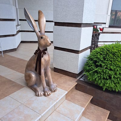 This image depicts a decorative statue of a rabbit, adorned with a brown velvet bow around its neck, positioned on a tiled outdoor staircase. The rabbit statue is light brown and appears to be made of a solid material. It is sitting upright with its paws resting on the steps. Behind the statue, a textured white pillar with dark brown horizontal accents is visible. To the right, a lush green bush is planted in a garden bed, bordered by small white pebbles and dark brown tiling. The surrounding architecture features white walls with similar dark brown accents. Red flowers in a pot are visible on a decorative metal railing in the background, near a window. The ground and steps are tiled with a combination of light beige and brown square tiles. The scene is set outdoors, suggesting a residential or public entrance area. The image is taken from a slightly low angle, looking up towards the statue and the building.