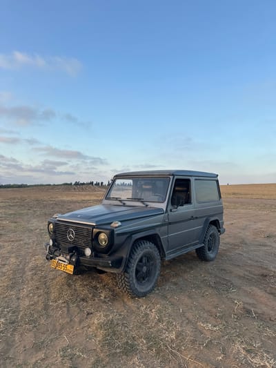 The image shows a gray Mercedes-Benz G-Wagon parked in a field of brown grass in Bnei Zion, Israel (latitude: 32.226, longitude: 34.849). The vehicle is facing the camera. The sky is blue with white clouds. There are no people in the image. The sun is low in the sky, casting long shadows.