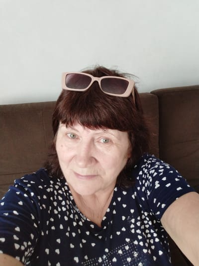 A middle-aged woman with dark brown hair and light green eyes is pictured from the chest up. She is wearing pink-framed sunglasses on top of her head, and a dark blue t-shirt with a white heart pattern. She is seated on a brown sofa against a plain white wall. The photo was taken in the city of Bezlesnyy, Russia.
