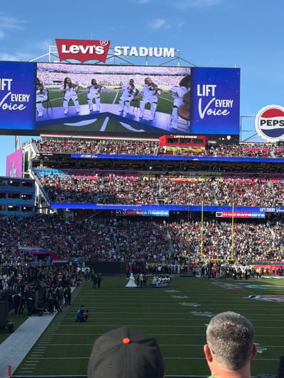 The image depicts a crowded stadium with a football field visible. On the left, a large screen displays a performance with the words "LIFT EVERY VOICE" visible. The stadium is branded with "Levi's Stadium" and a "Pepsi" logo. The score on the screen shows "47:12 SEATTLE HAWKS 25 NEW ENGLAND PATRIOTS." People are on the field, possibly performers or staff, with a photographer visible. A sea of spectators fills the stands.