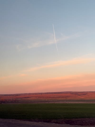 The image depicts a view of the landscape in Камаево, Russia, featuring a sky with hues of blue, white, and peach, possibly during sunrise or sunset. A faint contrail cuts across the blue portion of the sky. Below the sky, the landscape shows fields of green and brown, possibly farmland or natural terrain. The image appears to be taken from a high vantage point, slightly rotated. No people, specific objects, or interactions are visible.