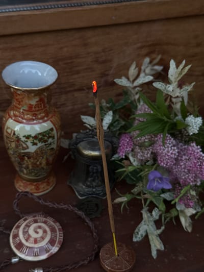 In Krásná Lípa, Czechia, a still life arrangement is captured on a wooden surface. An ornate vase, decorated with painted scenes of figures and foliage, stands to the left. Next to it, a small, dark, metallic object with intricate detailing is partially visible. In the foreground, a braided leather cord encircles a round pendant with a spiraling design in red and white. The central focus is an incense stick, glowing red at its tip and emitting a thin plume of smoke, resting in a circular brown holder. Behind these objects, a bouquet of flowers and foliage, featuring delicate pink and white blossoms, a single bluebell, and pale variegated leaves, adds a natural element to the composition. The background is a textured wooden surface, possibly a wall or furniture.