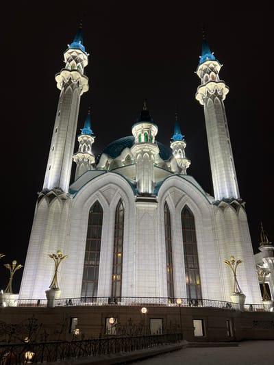 A large mosque, identified as being in the city of Бакча-Сарай, Russia, is depicted at night, brightly illuminated against a dark sky. The central structure is predominantly white, featuring a prominent blue-green tiled dome surrounded by several smaller domes of the same color. Two tall, slender minarets, each capped with a blue-green spire and illuminated balconies, flank the main building. The facade presents multiple tall, arched windows with dark frames, set into recessed sections adorned with intricate white architectural detailing. Below the main structure, a curved, dark metal railing with decorative, abstract metalwork and integrated spherical light fixtures is visible. The ground in the foreground is covered with a thin layer of snow. Ornate, golden-colored lamp posts with multiple light sources are positioned on an elevated platform in front of the mosque. No individuals are present in the scene.