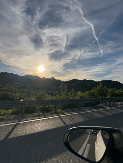 The image captures a scenic view from a car driving on a road near Očinići, Montenegro. The sun, partially obscured by wispy clouds, casts a warm glow over a landscape of rolling, tree-covered hills. The sky is a mix of blue and white, with streaky contrails from aircraft visible. A metal guardrail runs along the side of the road, separating it from the vegetated hillside. In the foreground, the car's side mirror reflects the road behind, showing another car and the white lane markings. The road itself is asphalt with white lines indicating lanes. The overall impression is of a peaceful, natural setting on a clear day, possibly late afternoon or early morning given the sun's angle.