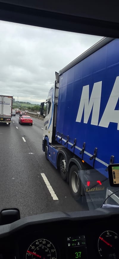 The image captures a scene on a highway, likely in the vicinity of Rotherham, United Kingdom, given the presence of a Scania truck and British license plates. The perspective is from the driver's seat of a vehicle, looking forward and slightly to the right. Traffic is heavy, with multiple vehicles visible on the multi-lane highway under an overcast sky.
Dominating the right side of the frame is a large blue truck with "MARITIME" written vertically on its side. The truck is a semi-trailer truck, with a distinct blue trailer and a white cab. The trailer has large white lettering spelling out "MA" (with the rest obscured by the cab). The license plate of the truck is visible as "AY73 YYP".
In front of the blue truck, other vehicles are seen, including a white truck with "PRIN Logistics" on its rear and a red sports car, likely an Audi R8, with the license plate "S644 UAY". The road surface is wet, suggesting recent rain. White lane markings are clearly visible.
The dashboard of the vehicle from which the photo is taken is partially visible at the bottom, showing a speedometer reading around 37 mph and a digital display indicating the time as 17:45.
The environment is a typical motorway landscape, with grassy verges and distant hills visible beyond the road. The overall impression is one of a congested but orderly flow of traffic on a main arterial route.