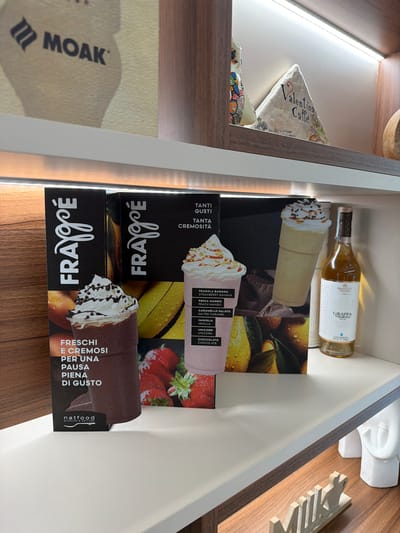 The image shows an eye-level, interior shot of a shelf or drawer.

Objects present include a bottle of "GRAPPA" liquor, along with several boxes of a product called "FRAYgoÉ" by "natfood", which appear to be dessert mixes or flavorings. The boxes feature images of frappes and list various flavors such as strawberry banana, peach mango, salted caramel, vanilla, unicorn, and chocolate.

Other items visible include a decorative dish labeled "Valentina dal Caffe" and a small white decorative sculpture.

The lighting in the shelf is provided by an LED strip.

The scene is likely in a kitchen or storage area, although the location cannot be pinpointed to Rosolini, Italy, with only the image data.