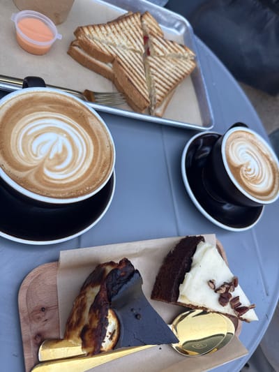 The scene depicts a grey table laden with food and beverages, located in Binyamina - Givat Ada, Israel. Two black ceramic coffee cups with white interiors and saucers are present; the larger cup in the foreground displays intricate latte art resembling a heart and rosetta pattern, while the smaller cup to the right features a simpler spiral design.
A rectangular metal tray, positioned on the upper left, holds four triangular halves of grilled sandwiches, characterized by distinct parallel grill marks. A red filling is visible in one sandwich, and a silver-colored fork rests beside them. Adjacent to the tray is a small, clear plastic cup containing an orange-hued sauce.
In the lower foreground, a wooden serving board is present, covered with brown parchment paper. On this paper, two distinct dessert items are arranged. To the left, a slice of Basque cheesecake exhibits a heavily charred, dark brown to black exterior, contrasting with a lighter, creamy interior. A gold-colored serving utensil, possibly a spoon or spatula, is partially inserted into this dessert. To the right, a slice of dark brown cake, presumably chocolate or carrot, is topped with a thick layer of white frosting and garnished with several pecan halves. Another gold-colored spoon is placed alongside this cake.
No individuals are visible in the image, but the arrangement of items suggests a prepared setting for consumption. The lighting indicates an outdoor or brightly lit indoor environment.