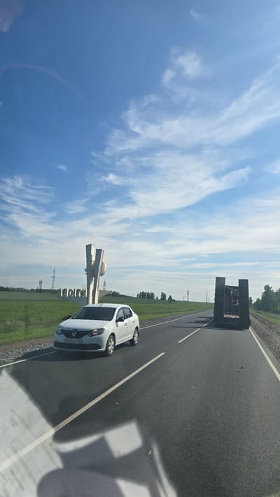 The image depicts a highway scene under a partly cloudy blue sky. On the left side of the road, a white sedan is parked. In the background, there is a large concrete structure with Cyrillic lettering that reads "Актюбинский," indicating the location is Aktubinsky, Russia. Further down the road, a flatbed trailer carrying heavy equipment is visible. The environment is rural, with green fields and trees lining the road. The perspective suggests the photo was taken from another vehicle moving on the highway.