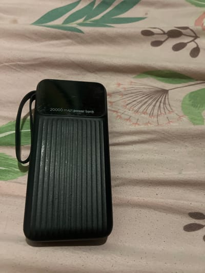 A black power bank with a capacity of 20000 mAh is lying on a pink and white patterned fabric. The power bank has a textured surface with vertical lines and a smooth top section with text. A black strap is attached to the left side of the power bank. The fabric has a floral or leafy pattern with green and pink elements. The image appears to be taken indoors, likely on a bed or sofa. There is no indication of the location being in the city of Astoņi, Latvia.