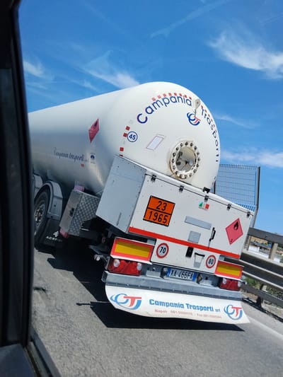 The image depicts the rear-right side of a white tank semi-trailer, observed from a slightly elevated perspective on an asphalt highway. The sky is bright blue with scattered cirrus clouds.
The semi-trailer's cylindrical tank is white, bearing the company name "Campania Trasporti s.r.l." in cursive and "Campania Trasporti" in larger, curved blue and red lettering with a stylized "CT" logo. A white circle containing "45" is visible on the tank. A red diamond placard with a flame symbol and the numeral "2" indicates flammable gas.
The rear structure of the trailer features an orange hazard panel displaying the numbers "23" above "1965", which corresponds to UN 1965 (Liquefied Petroleum Gas) and its hazard identification number. An Italian flag sticker with "MADE IN ITALY" is present. Another red diamond placard (flame, '2') is affixed to the right. Red circular speed limit signs showing "70" and "80" km/h are mounted on the rear bumper. The Italian vehicle registration plate reads "XA 466FA". A white mud flap at the bottom lists "Campania Trasporti srl", contact information including "Napoli 081-5590570 - Fax 081-5595084", and the "CT" logo. The brand "ACERBI" is visible on a mudguard.
No individuals are visible in the image. The vehicle is in motion on a paved road. In the background, a metal mesh fence and a concrete barrier run parallel to the road, with indistinct distant structures and sparse foliage beyond. The scene is consistent with a highway environment used for intercity transport, such as roads leading into or through Bisceglie, Italy. However, no specific visual landmarks within the image definitively identify the precise location within Bisceglie.