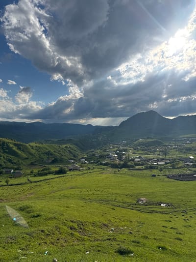 This is a vertical, high-angle shot of a mountainous rural landscape in Верхняя Мара, Russia. The sun is shining brightly, breaking through dramatic, partly cloudy skies on the left side of the image, casting rays of light. The right side of the image shows a verdant green valley dotted with houses, some with visible roofs, and a winding road. The valley slopes upwards towards a large, dark, and imposing mountain range that stretches across the horizon. The overall impression is one of a vast, natural landscape with human settlements integrated into it. The time of day appears to be daytime, likely late morning or early afternoon, given the strong sunlight. The weather is fair with scattered clouds. The scene evokes a sense of peacefulness and natural beauty.
