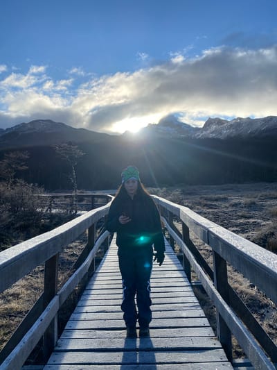 A photograph taken in Unknown, Argentina, depicts a woman standing on a wooden bridge. The bridge appears to be lightly frosted. The woman is wearing a dark jacket, dark pants with a light stripe down the side, and a green patterned hat. She is holding an object in her right hand. The sun is behind her, creating a lens flare effect on the right side of her body and partially obscuring the view. Mountains are visible in the background, partially obscured by clouds and the bright sunlight. The sky is blue with scattered clouds. The landscape around the bridge appears to be covered in dry grass or vegetation. The photo is taken at a slight angle, making the bridge appear diagonal.