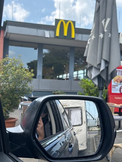 The image shows a view of a McDonald's restaurant in Frechen, Germany, reflected in a car's side mirror. In the reflection, a light-colored vehicle is visible behind the car. The exterior of the McDonald's building features a yellow "M" logo, large glass windows, and a gray awning with white stars. Green trees and foliage are also visible in front of the building. A red object is positioned near the awning. A human hand is partially visible in the car's side mirror.