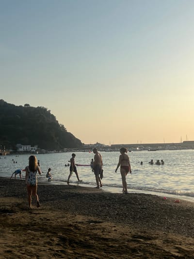 The image captures an evening scene on the dark sand and pebble beach of Baia del Silenzio in Sestri Levante, Italy, facing east towards the rocky promontory and the town's small harbor. The sky displays a clear gradient from light blue overhead to a warm yellow-orange near the horizon, indicative of late afternoon or early evening light.
Approximately 15-20 individuals are present. In the foreground, a young child in a patterned one-piece swimsuit walks away from the viewer towards the waterline on the left. Further left, another child in blue swim trunks performs a handstand or somersault at the water's edge, near a third small child seated in the shallow water. Moving right along the waterline, a pre-teen boy in dark swim trunks walks leftward holding a light-colored float. Beside him, an adult male, bald or closely shaven, in dark swim trunks, walks leftward carrying a dark object. To the far right in the foreground, an adult female in a dark red two-piece bikini walks away from the viewer towards the water, holding a lit cigarette in her right hand.
Several other individuals are visible in the mid-ground water, partially submerged, swimming or wading. The calm water of the bay extends to a distant shoreline on the right, where numerous small to medium-sized boats are moored at a marina or pier. White buildings are discernible at the base of a steep, densely vegetated hill that dominates the left background. A prominent single tree stands atop the hill. Two small, colorful plastic beach toys (red and pink) are visible on the dark sand near the waterline in the far right foreground.