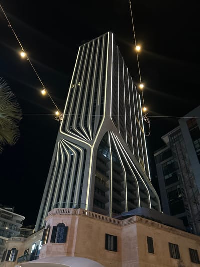 A striking nighttime view showcases a modern building illuminated with horizontal lines of light, its unique design curving inward towards the middle. Adjacent to it is a more traditional structure with a beige facade and rectangular windows. String lights hang diagonally across the scene, adding to the nighttime ambiance. The composition is shot from a low angle, with dark sky filling the negative space. A palm tree is at the bottom of the frame.