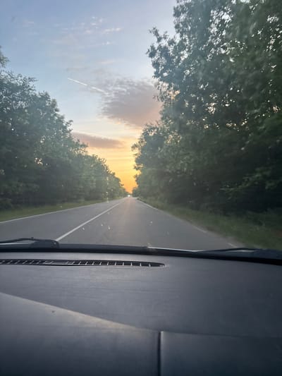 The image shows a view from inside a vehicle, likely a car, driving along a paved road. Dense trees line both sides of the road, creating a tunnel-like effect. In the distance, the setting sun is visible, casting a warm glow. The dashboard of the car is visible in the foreground, occupying the right side of the frame. There is a contrail visible in the sky. The image is rotated counter-clockwise by 90 degrees. The location is identified as Stolnik, Bulgaria.
