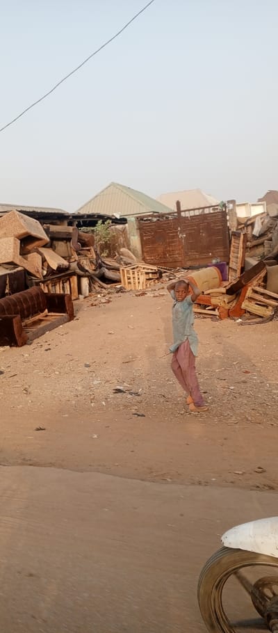 The image depicts an outdoor scene in Tunga Mekidi, Nigeria, characterized by significant clutter and debris.
**Person:** A young individual, appearing pre-teen, stands slightly right of the frame's center on a dirt path. They wear a light blue or grey long-sleeved garment and reddish-brown or pink baggy trousers, with brown sandals on their feet. Their hands are positioned behind their head, and they maintain a direct, neutral gaze towards the camera.
**Environment:** The ground consists of uneven, light brown dirt or sand, scattered with small pieces of debris. To the left and extending across the mid-ground, large, disorganized piles of discarded materials are evident. These piles primarily comprise broken furniture, including sofa frames (some with remnants of upholstery, others stripped) and numerous wooden pallets or crates. A brown sofa frame with exposed wooden supports is prominent in the lower-left corner. In the background, several buildings with corrugated metal roofs are visible, one featuring a light green roof and another a lighter, possibly white or grey, roof. A large, heavily rusted metal gate with a latch mechanism is situated in the mid-background, to the right of the central debris piles. The sky is clear, pale blue, or light grey. A single thin, dark wire crosses the upper-left portion of the sky.
**Objects/Actions:** The young person is standing in a casual pose. In the immediate foreground, at the bottom right, a portion of a white fender or fairing and the front wheel of a vehicle, possibly a motorcycle or bicycle, are partially visible, suggesting the photograph was taken from a vehicle. No direct interactions are depicted. The overall scene indicates an area used for the storage or processing of discarded materials, resembling a salvage yard or informal waste collection site.