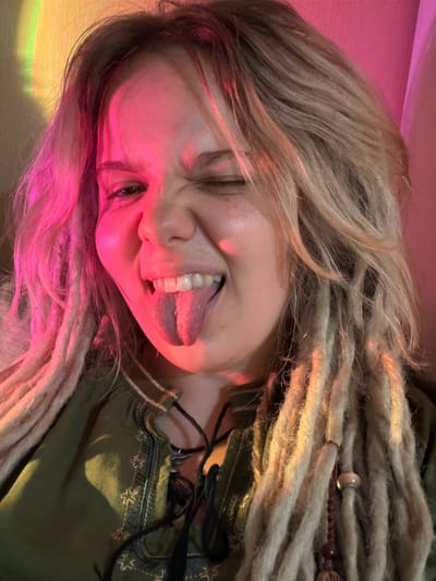 The image shows a close-up selfie of a young woman with blonde dreadlocks and freckles. She is winking and sticking out her tongue, which appears to have a darker purple tint on the tip. She is wearing a green, embroidered top. The lighting in the image is dramatic, with pink and yellow hues cast across her face and hair, possibly from a light effect or colored lighting. The background is blurred and appears to be a wall with some shadowy areas. There are no discernible elements that would place the location specifically within Vostochny District, Russia.