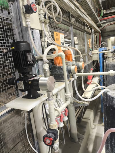 The image shows a section of an industrial chemical dosing system in Dimona, Israel. The system consists of pumps, valves, tubing, and tanks, all arranged in a functional but somewhat disorganized manner. A black electric motor is visible, connected to a pump with a stainless-steel head. Several transparent tubes carry fluids, connecting various components. Red-handled valves are present, indicating manual control points. A pressure gauge is mounted on a section of piping. In the background, metal grating and shelving are visible, as well as some warning signs. The overall environment appears to be an indoor industrial facility, possibly a water treatment plant or a chemical processing facility, given the nature of the equipment. The lighting suggests it is daytime, but the specific source is not clear. There are no people visible in the image. The area appears to be dusty and shows signs of chemical residue.