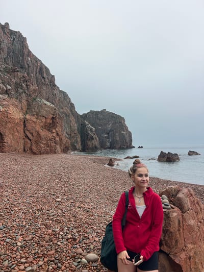 A young woman stands on a pebble beach in Russkiy Island, Russia, against a backdrop of rugged red rock cliffs and the sea under an overcast sky. She wears a red zip-up jacket over a white top, black shorts, and has a backpack over one shoulder. Her hair is pulled back in a bun, and she is holding a smartphone. The beach is made up of small, reddish-brown pebbles, and a large rock formation is visible in the background near the shoreline. The sea is calm with scattered rocks breaking the surface. The sky is a uniform grey.
