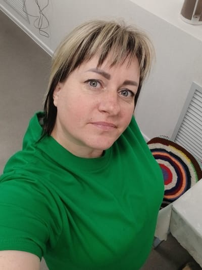 The image is a medium shot selfie of a woman with blonde and dark brown hair, and blue eyes. She is wearing a green hoodie and looking at the camera. In the background, there is a wall with some wires, a louvered vent, and a colorful crocheted circular rug on a table covered with a white tablecloth. The scene appears to be indoors, possibly in a residential setting in Prokudskoye, Russia.