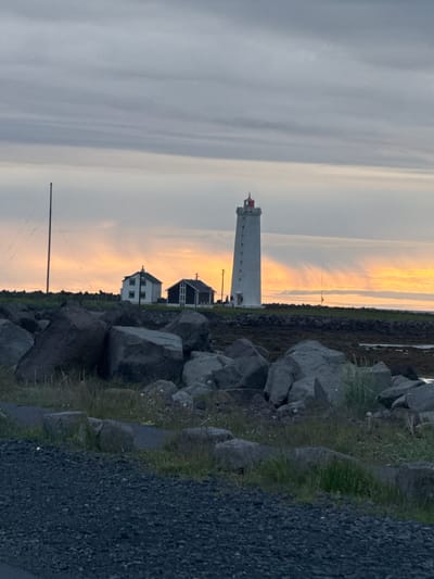 A lighthouse stands on a rocky shoreline in Seltjarnarnesbær, Iceland, under a cloudy sky. The lighthouse is tall and white with a red top. Two small buildings are visible to the left of the lighthouse, one white and the other dark. The foreground is filled with large rocks and some green grass. The background shows a vibrant orange and yellow sunset peeking through the clouds.