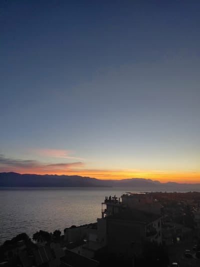 The image captures a twilight scene overlooking the Municipal Unit of Aegio, Greece. In the foreground, dark silhouettes of buildings, some with visible rooftops and windows, dominate the lower portion of the frame. Trees with their dark foliage are also present. A body of water, likely the Gulf of Corinth, occupies the mid-ground, its surface reflecting the soft hues of the sunset. Beyond the water, a range of mountains forms a dark, jagged horizon. The sky above transitions from a deep blue at the top to warmer tones of orange, pink, and yellow near the horizon, indicating either sunrise or sunset. The overall impression is one of a tranquil evening or early morning view of the coastal city.