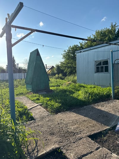 The image captures a rural scene in the village of Verkhneyarkeyevo, Russia, during daylight. In the foreground, a concrete pathway is visible, partially covered by green grass and some leafy plants. To the left, a vertical blue pole supports a wire that extends horizontally across the frame. Behind the pathway, a tall, green, A-frame structure stands prominently in a grassy area. Further back, a corrugated metal building with a small, multi-paned window is situated to the right. Lush green trees and vegetation surround the structures. The sky above is blue with scattered white clouds, and the sun is shining, casting shadows. The overall impression is one of a functional, outdoor space in a somewhat rustic setting.