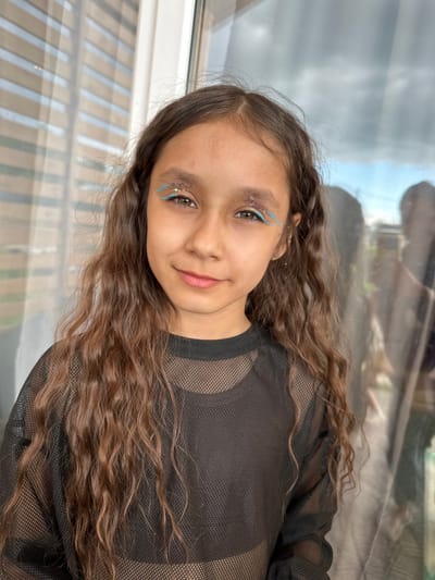 A young girl with long, wavy brown hair and brown eyes is shown in a medium shot, looking directly at the camera. She has on a black, mesh long-sleeved shirt. Her face is adorned with light blue winged eyeliner and glitter around her eyes. She is standing in front of a window with horizontal blinds, and a reflection of trees and the sky can be seen through the glass. The background is softly blurred, creating a shallow depth of field. The lighting is natural and appears to be from the side.